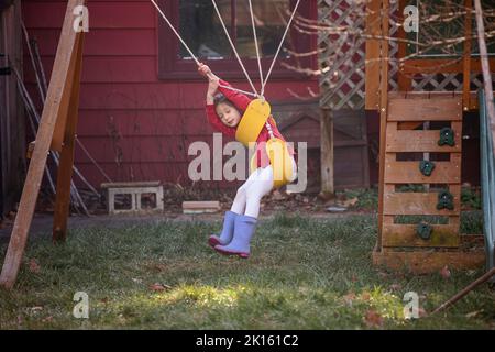 Una bambina con stivali da pioggia si siede felicemente sulle altalene aggrovigliate Foto Stock