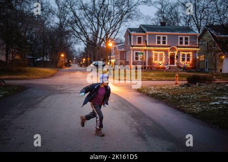 Un ragazzo nei vestiti di inverno gioca sulla strada di lamplit al crepuscolo Foto Stock