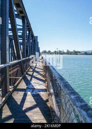 Conduce via passerella attraverso il ponte ferroviario storico con modelli di camion in ombra sul porto di Tauranga. Foto Stock