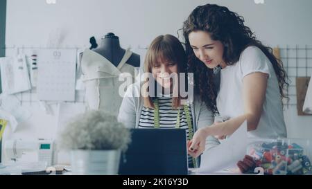 I designer di abbigliamento allegri e creativi guardano lo schermo del notebook, conversano in modo amichevole, gestendo, sorridendo e ridendo insieme in un laboratorio leggero. Concetto di divertimento sul posto di lavoro. Foto Stock