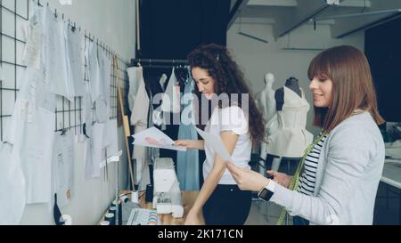 Attractive women clothing designers looking at garment sketches hanging on wall, discussing them, choosing new images for newest collection. Friendly informal atmosphere in studio. Foto Stock