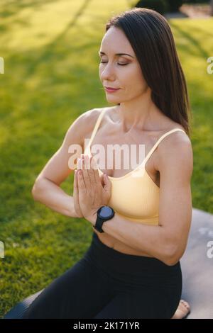 Ritratto di una donna seduta sul materassino di idoneità che fa l'esercitazione di yoga nel parco pubblico verde. Il concetto di calma e meditazione. Meditazione. Yoga in Foto Stock