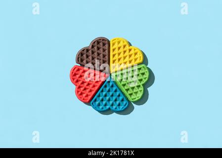 Vista dall'alto con cialde a forma di cuore multicolore in luce brillante su sfondo blu. Waffle arcobaleno disposti minimalista su un tavolo colorato. Foto Stock