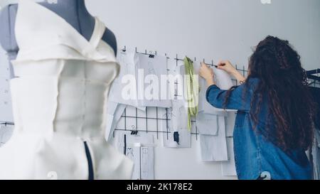 Giovane donna designer di abbigliamento sta guardando schizzi e disegni appesi alla parete in un laboratorio leggero. Grande collezione di bozze sopra il banco di sartoria, manichino vestito in primo piano. Foto Stock