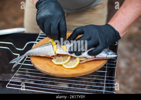 Sgombro alla griglia con fettine di limone, vista dall'alto. Il processo di cottura del pesce all'aperto. Chef in guanti in lattice nero preparazione di sgombro pesce su un bosco Foto Stock