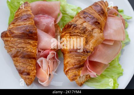 Croissant sandwich con prosciutto (jamon, prosciutto crudo) e lattuga. Snack deliziosi, antipasti, tapas. Prima colazione. Fast food per tr Foto Stock