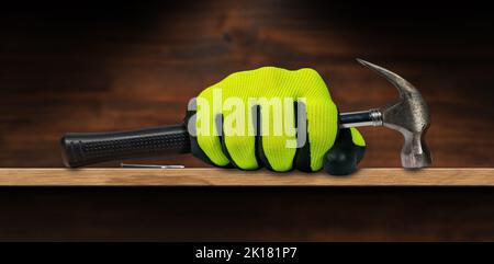 Mano con guanto di protezione che tiene un vecchio martello a artiglio in acciaio con manico in gomma nera, su un banco di lavoro in legno con spazio per unghie e fotocopie. Foto Stock