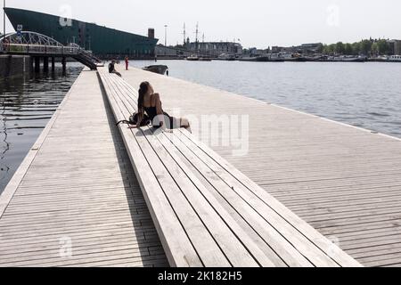 Una giovane donna in un breve vestito nero siede al sole su un pontile vicino a un canale in una giornata calda e soleggiata ad Amsterdam, nei Paesi Bassi. Foto Stock