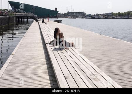 Una donna in un abito nero corto siede al sole su un pontile vicino a un canale in una giornata calda e soleggiata ad Amsterdam, nei Paesi Bassi. Foto Stock