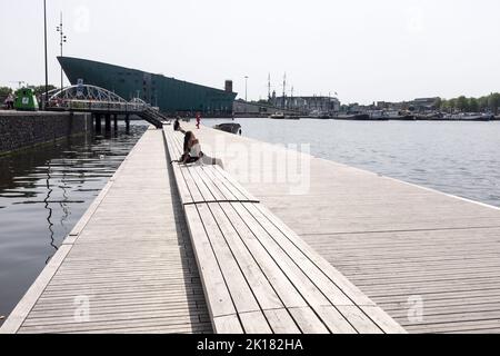 Una giovane donna con un breve vestito nero siede al sole su un pontile vicino a un canale in una giornata calda e soleggiata ad Amsterdam, nei Paesi Bassi. Foto Stock