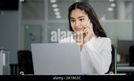 Giovane donna araba si siede in ufficio colloqui sul telefono guarda schermo laptop rende ordine in negozio online ordini consegna businesswoman consulente remoto Foto Stock