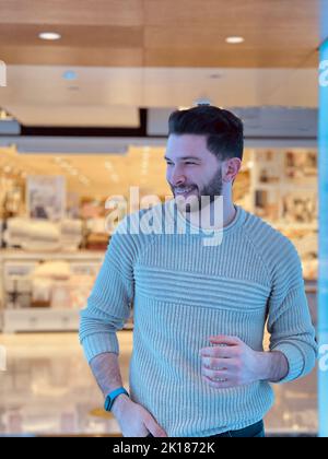 Un uomo che indossa un maglione azzurro sorridente, in piedi in un centro commerciale con piastrelle bianche Foto Stock