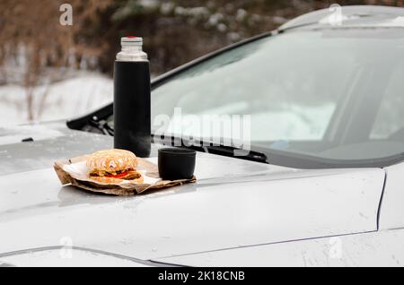 Picnic invernale sul lato della strada. Hamburger e thermos con caffè sul cappuccio della macchina Foto Stock