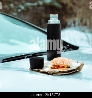 Picnic invernale sul lato della strada. Hamburger e thermos con caffè sul cappuccio della macchina Foto Stock