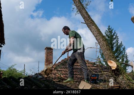 Un uomo pulisce il tetto di una casa dopo una grande tempesta in un giorno d'estate. Un uomo si alza sul tetto e rimuove le foglie e i rami caduti Foto Stock