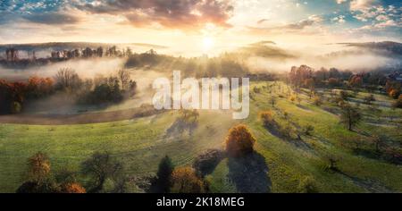 Magnifico scenario di alba aerea che mostra un paesaggio rurale, con prati, campi, colline, alberi, e bei raggi solari che cadono attraverso la nebbia Foto Stock