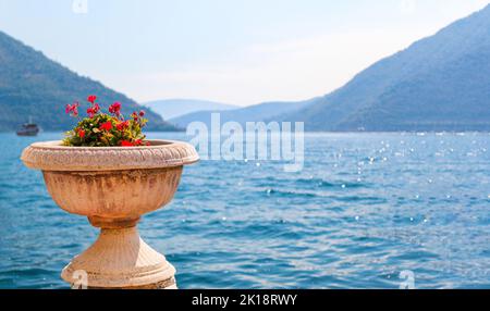 Fiori rossi in vaso di pietra d'epoca con bel mare Adriatico blu e montagne. Concetto estivo tropicale con spazio copia. Perast, Montenegro Foto Stock