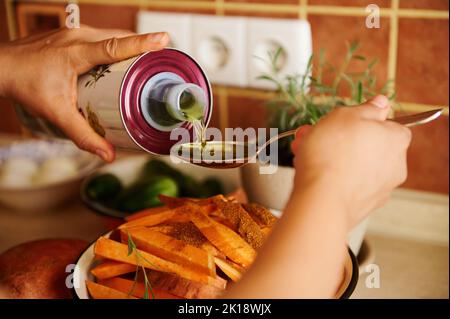 Le mani dello chef versano l'olio d'oliva biologico su un cucchiaio e lo versano sulle fette di patate dolci biologiche mentre preparano il pranzo Foto Stock