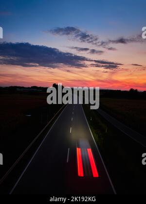 Uno scenario drammatico di un tramonto vibrante nel cielo con un'auto che guida su una strada asfaltata Foto Stock
