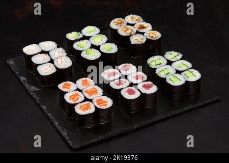 Assortimento di diversi tipi di sushi a rullo posti su tavola di pietra nera. Sushi con salmone, cetrioli, formaggio spalmabile, avocado e semi di sesamo Foto Stock
