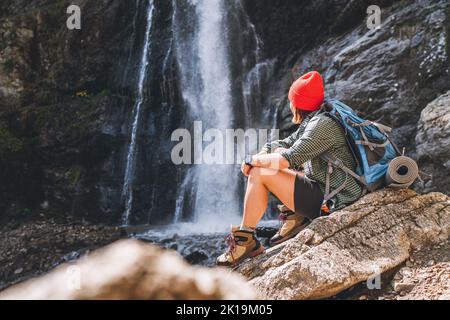 Donna con uno zaino in cappello rosso vestito con abiti da trekking attivi seduta vicino alla cascata del fiume montagna e godendo del potere della natura. Foto Stock
