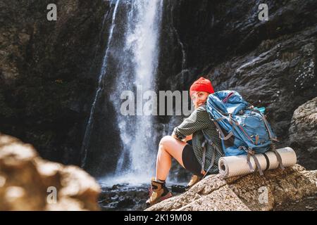 Donna sorridente con zaino in cappello rosso vestito con abiti da trekking attivi e stivali seduti vicino alla cascata del fiume di montagna e godersi gli spruzzi Natur Foto Stock