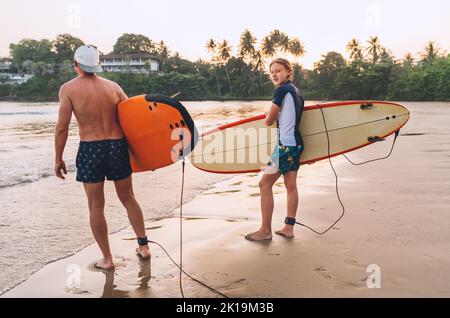 Ragazzo con padre e tavola da surf va in mare per fare surf sull'isola dello Sri Lanka. Hanno una vacanza invernale e godono di una bella luce al tramonto Foto Stock