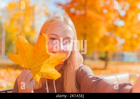 studentessa bionda che prende un selfie e tiene una foglia autunnale sullo sfondo di alberi autunnali gialli e sorridente. Foto Stock
