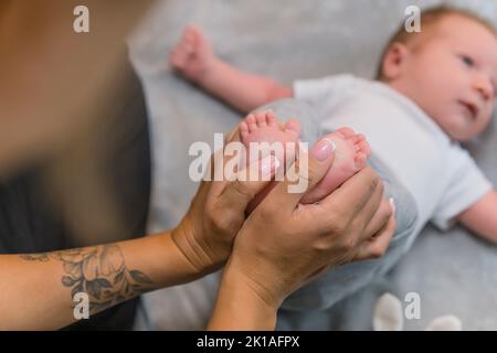 Una nuova madre amorevole che tiene delicatamente i suoi figli piccoli piedi. Un ragazzino dai capelli castani carini, adagiato sulla schiena su una coperta grigia con le braccia tese ai lati. Foto di alta qualità Foto Stock