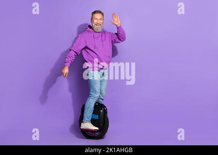 Foto di bel pensionato uomo funky senior vestito porpora felpa con cappuccio guida monowheel ondulazione Palm saluti isolati su sfondo di colore viola Foto Stock