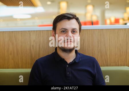 guardando la macchina fotografica sorridente giovane uomo della barba caucasica non rasata di 32 anni in una polo blu marina in un bar presso la corte alimentare. In attesa di concetto, lui Foto Stock