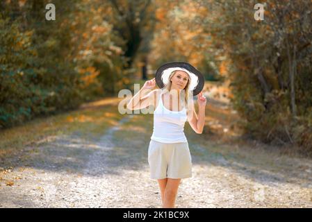 Bella donna bionda sorridente amichevole indossando un cappello estivo bianco e nero in una parte superiore bianca con le mani sul cappello contro un bel bac autunnale Foto Stock