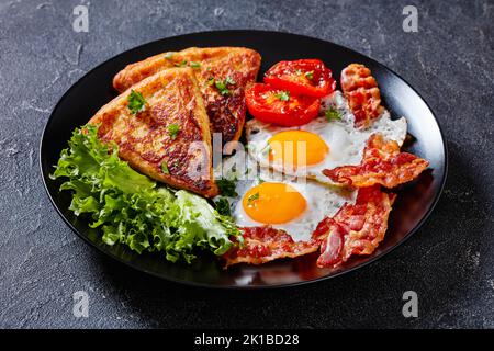 colazione irlandese con uova fritte, fette di rasher o pancetta, pane di patate fritto, pomodori alla griglia e lattuga fresca su piatto nero su tavolo di cemento Foto Stock