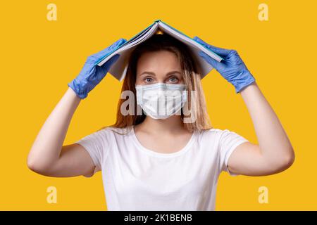 Istruzione in quarantena. Distanza sociale. Studentessa insicura nei guanti per maschera facciale che proteggono la testa con libro aperto sul giallo brillante. Coronav Foto Stock