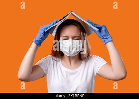 Coronavirus panico. Rischio di infezione. Studentessa ansiosa in maschera guanti di protezione della testa coprendo con libro aperto isolato su arancione. Quarantena h Foto Stock