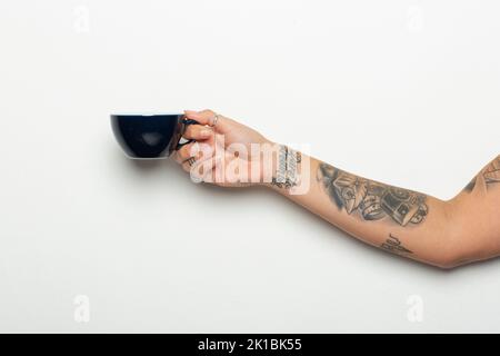 Colpo di primo piano di un braccio e di una mano di una femmina tatuata con una tazza di caffè su sfondo bianco Foto Stock