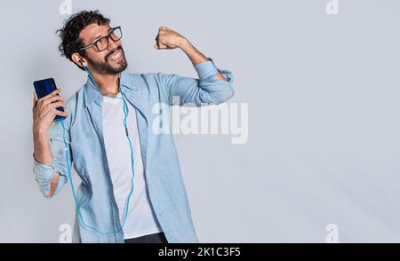 Felice uomo bello con le cuffie che tengono un telefono cellulare e celebrare, felice persona utilizzando il suo smartphone e celebrare la vittoria isolato Foto Stock