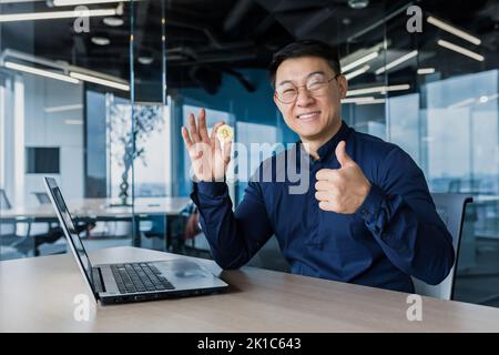 Uomo d'affari asiatico di successo sorridente e guardando la macchina fotografica, tenendo la moneta bitcoin e dando pollici in su, uomo che lavora all'interno di un moderno edificio di uffici utilizzando un computer portatile, ha rotto la borsa Foto Stock