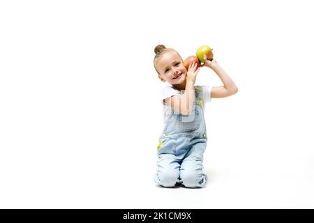 Ragazza carina vestita con tute in denim che girano con mele su sfondo bianco. Ragazza che tiene le mele per le orecchie Foto Stock