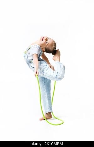 Danzatrice flessibile piccola ginnastica acrobat ragazza stretching corpo, mettere i piedi sulla testa. Bambina vestita con tuta in denim con corda da salto. Foto Stock