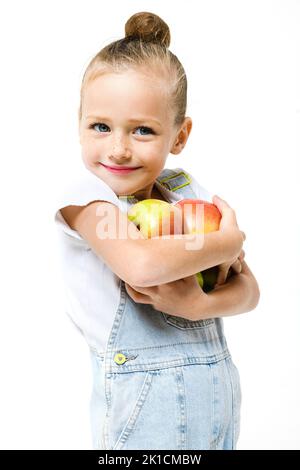 Ragazza carina vestita con tute in denim che reggono un'armatura di mele fresche su uno sfondo bianco. Foto Stock