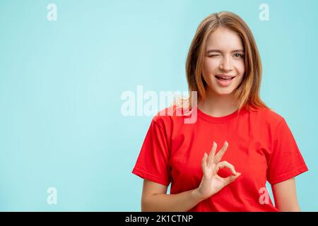 Gesto OK. Scelta perfetta. Ritratto di entusiasta donna positiva in t-shirt rossa che accetta l'offerta winking isolato su copia blu spazio commerciale indietro Foto Stock
