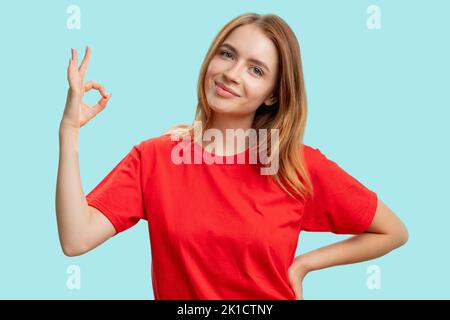 Segnale OK. Bravo. Ritratto di donna amichevole soddisfatto in t-shirt rossa incoraggiando con approvazione mano gesto sorridente isolato su sfondo blu. De Foto Stock