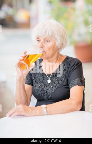 Donna matura seduta a un tavolo in un bar estivo e bere birra Foto Stock