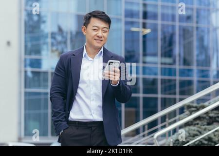 Riuscito uomo d'affari asiatico nel vestito di affari che cammina fuori vicino all'edificio dell'ufficio, direttore maschio del direttore usando il telefono, sorridente e felice leggendo il messaggio in linea. Foto Stock