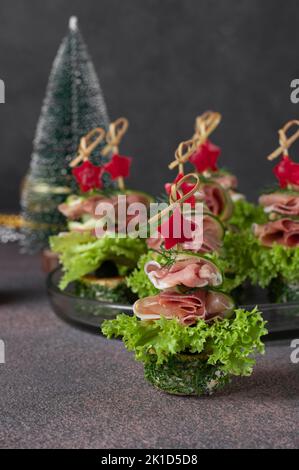 Festive tartine in forma di alberi di Natale da prosciutto, cetrioli, foglie di lattuga e stelle di peperone su sfondo marrone Foto Stock