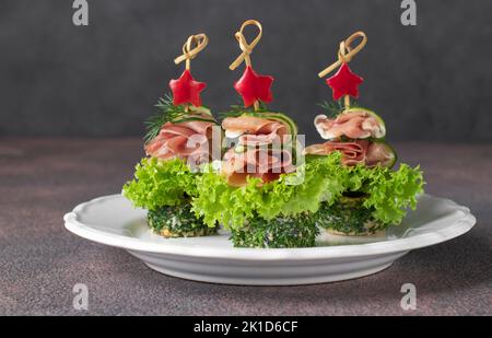 Festive tartine in forma di alberi di Natale da prosciutto, cetrioli, foglie di lattuga e stelle di peperone su piatto bianco su sfondo marrone Foto Stock