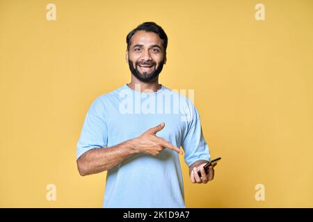 Uomo indiano felice che indica il telefono cellulare isolato su sfondo giallo. Foto Stock