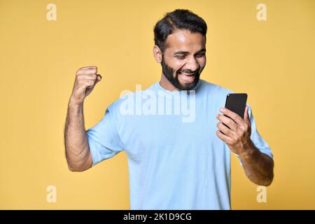 Uomo indiano eccitato che alza il pugno utilizzando smartphone isolato su sfondo giallo. Foto Stock