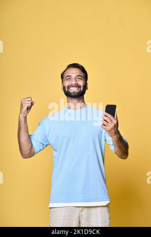 Uomo indiano eccitato che alza il pugno utilizzando telefono isolato su giallo, verticale. Foto Stock